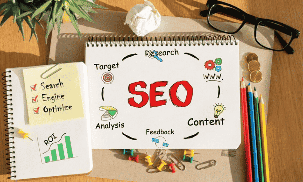A notebook page with "SEO" in bold letters surrounded by related terms, next to sticky notes with "Search Engine Optimize" and a graph indicating ROI, pencils, pushpins, and eyeglasses on a desk.