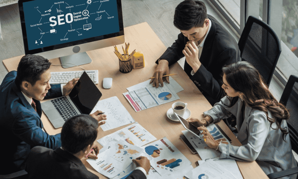 Four professionals are gathered around a conference table reviewing SEO data on papers and laptops, with a large monitor displaying SEO-related terms.